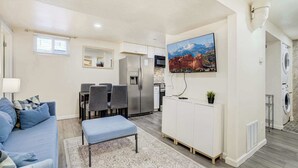 Un espacio habitable compacto con kitchenette, zona de comedor, sala de estar con sofá y reposapiés, y televisor montado.