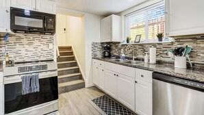 Una cocina moderna con armarios blancos, lavavajillas de acero inoxidable, microondas y una ventana con persianas.