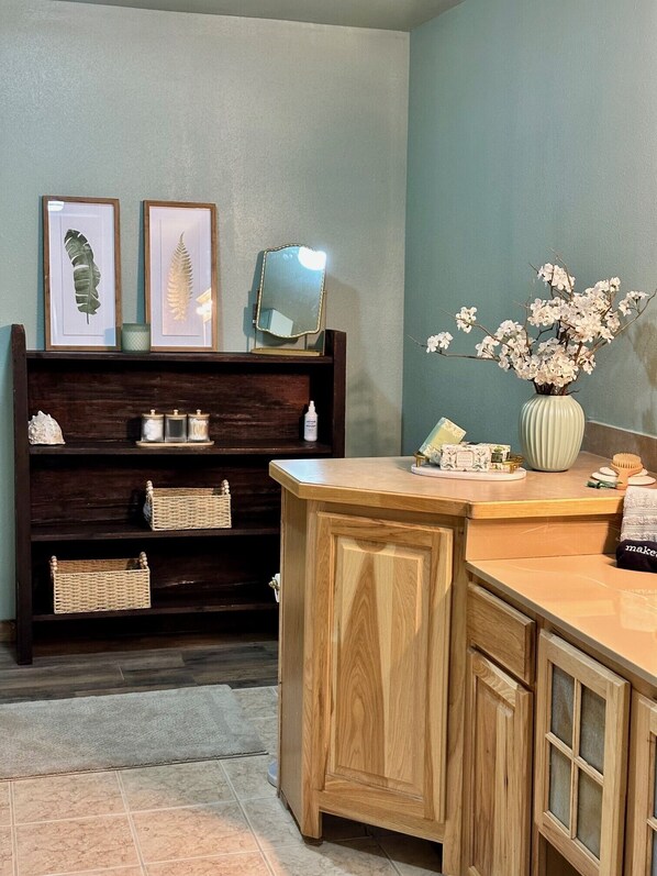 A bathroom with a wooden vanity, a shelf with framed botanical prints, a mirror, and a vase with flowers.