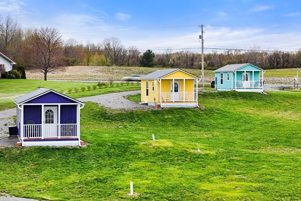 Three small, single-story houses with distinct colors and simple designs, situated on a grassy slope with a clear sky.