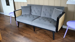 Un canapé gris à motifs, doté de pieds noirs et d’accoudoirs en osier, placé entre deux petites tables violettes sur un plancher en bois.