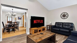 Una sala de estar con televisor mostrando 'NETFLIX', un comedor con mesa y sillas de madera, un sofá de cuero negro y un elemento decorativo en la pared.
