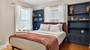 Un dormitorio con cama, ventilador de techo, estantería y ventana con cortinas.