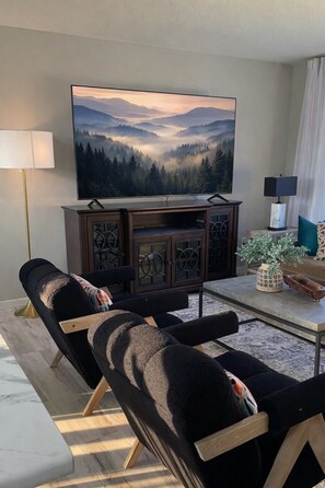 Un salon équipé d’un téléviseur à écran plat, d’un meuble en bois, de deux canapés noirs, d’une table basse et d’un lampadaire.