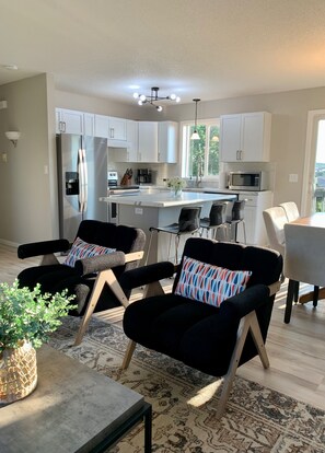 Une cuisine moderne avec des meubles blancs, un îlot central et un coin repas comprenant des chaises noires et un tapis à motifs.