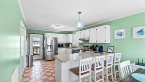 A kitchen with white cabinets, a stainless steel refrigerator, a granite countertop, and a bar area with white chairs. - "Dibbs on Holden" 3 Bedroom, 3 Bathroom, Canal Home (Holden Beach)