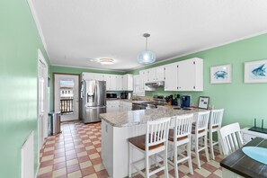 A kitchen with white cabinets, a stainless steel refrigerator, a granite countertop, and a bar area with white chairs. - "Dibbs on Holden" 3 Bedroom, 3 Bathroom, Canal Home (Holden Beach)