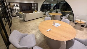 Et moderne mødelokale med en stor, buet glasvæg, grå polstrede stole og et centralt træbord med dokumenter.