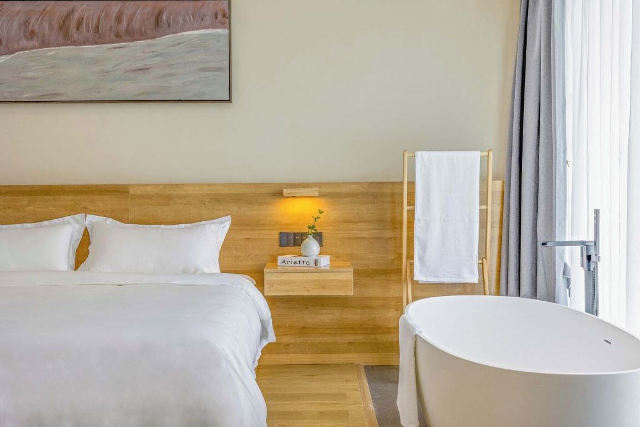 Une chambre d’hôtel avec une tête de lit en bois, un lit avec une literie blanche, une table de chevet ornée d’un vase et d’une plaque nominative, une baignoire blanche et un tableau accroché au mur.