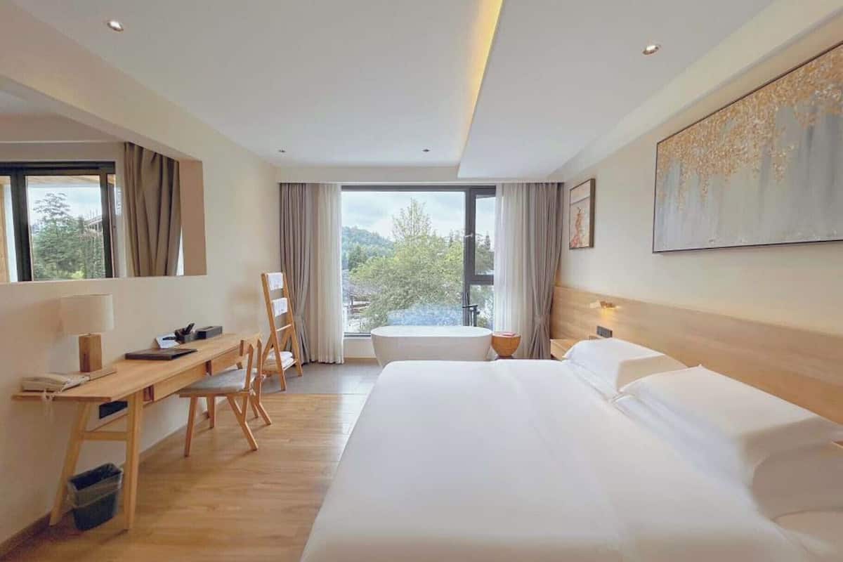 A hotel room with a large bed, a desk, a chair, a window with a view, and a painting on the wall.