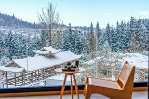 Quarto com vista para árvores e edifícios cobertos de neve, um banco de madeira e uma pequena mesa com um objeto decorativo.