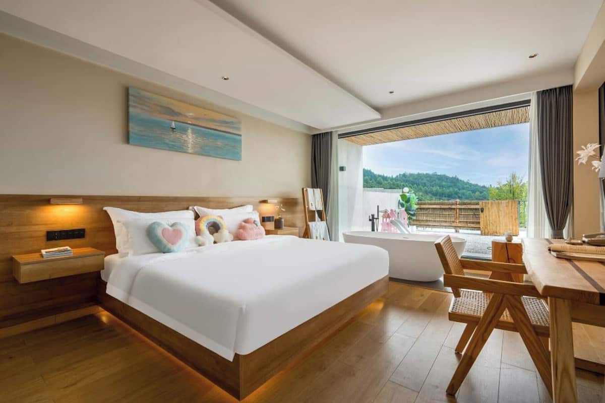 A modern bedroom with a large bed, a wooden nightstand, a desk with a chair, a bathtub, and a view of a green landscape.