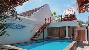 A swimming pool with a wooden staircase leading to a deck, surrounded by a white building with a tiled roof. - Casa Kai (Gili Trawangan)