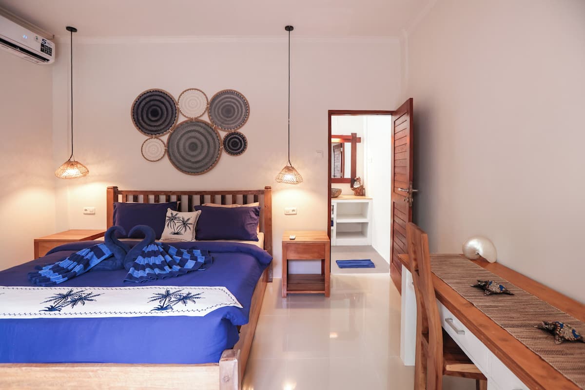 Un dormitorio con cama, mesitas de noche, un banco de madera, decoración en la pared y una puerta que da a otra habitación.