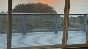 Ein Zimmer mit großem Fenster und Blick auf einen Sonnenuntergang über dem Meer, einem Balkon mit Geländer und einer Klimaanlage.