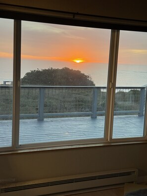 A room with a large window overlooking a sunset over the ocean, a balcony with a railing, and an air conditioning unit. - NEW LISTING! Oceanfront Coastal Home-SPECTACULAR VIEWS with Hot Tub & Large Deck (Gualala)