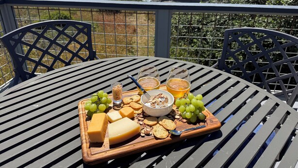 Uma mesa com uma tábua de queijos, uvas e bebidas em uma varanda com vista para o oceano.