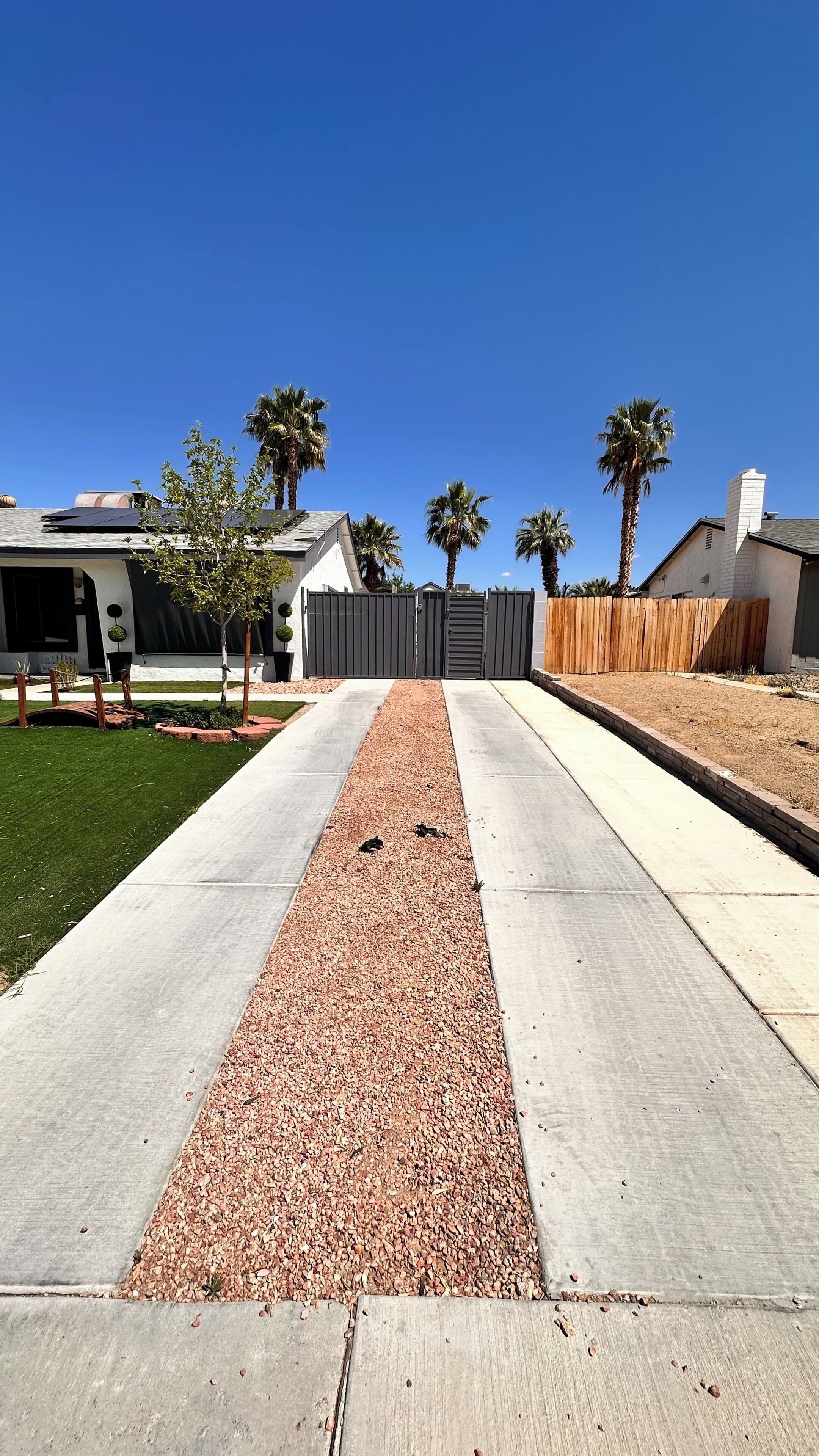 A residential driveway with a gravel path, palm trees, and a gated entrance.