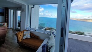 A room with a large window overlooking the sea, a sofa, and a small table. - Ocean edge, beach side holiday cottage in Sennen Cove with 180’ panoramic Views (Sennen Cove)
