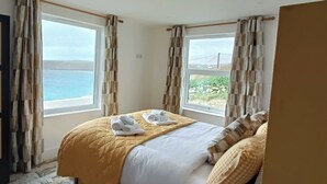 A hotel room with a bed, two windows, and a view of the sea. - Ocean edge, beach side holiday cottage in Sennen Cove with 180’ panoramic Views (Sennen Cove)
