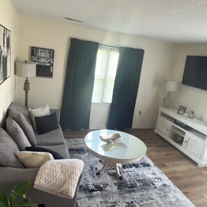 A living room with a sofa, a coffee table, a TV, and a window with curtains. - Charming 2-bedroom apartment within walking distance to beach (Myrtle Beach)