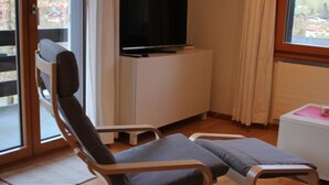 A room with a wooden floor, a television on a white cabinet, a window with curtains, and a chair with a cushion. - Ski-in/ Ski-out Nordostwand 4-Bett-Ferienwohnung by Interhome (Grindelwald)
