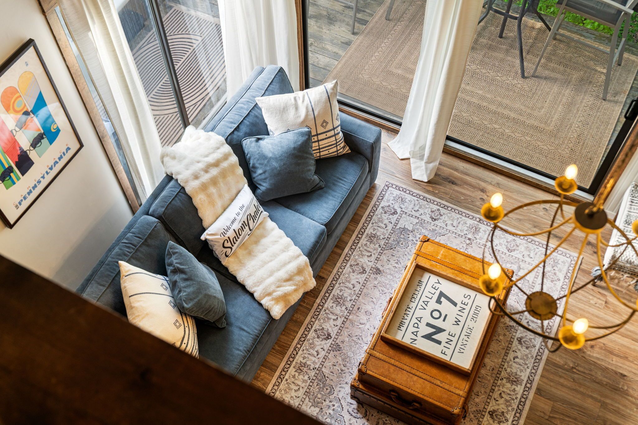 Un salon moderne avec un canapé bleu, des coussins décoratifs, un plaid blanc, un panneau d’accueil encadré, une vieille boîte à vin et un tapis à motifs.