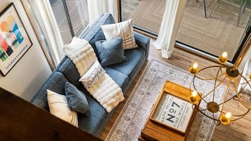 Un salon moderne avec un canapé bleu, des coussins décoratifs, un plaid blanc, un panneau d’accueil encadré, une vieille boîte à vin et un tapis à motifs.