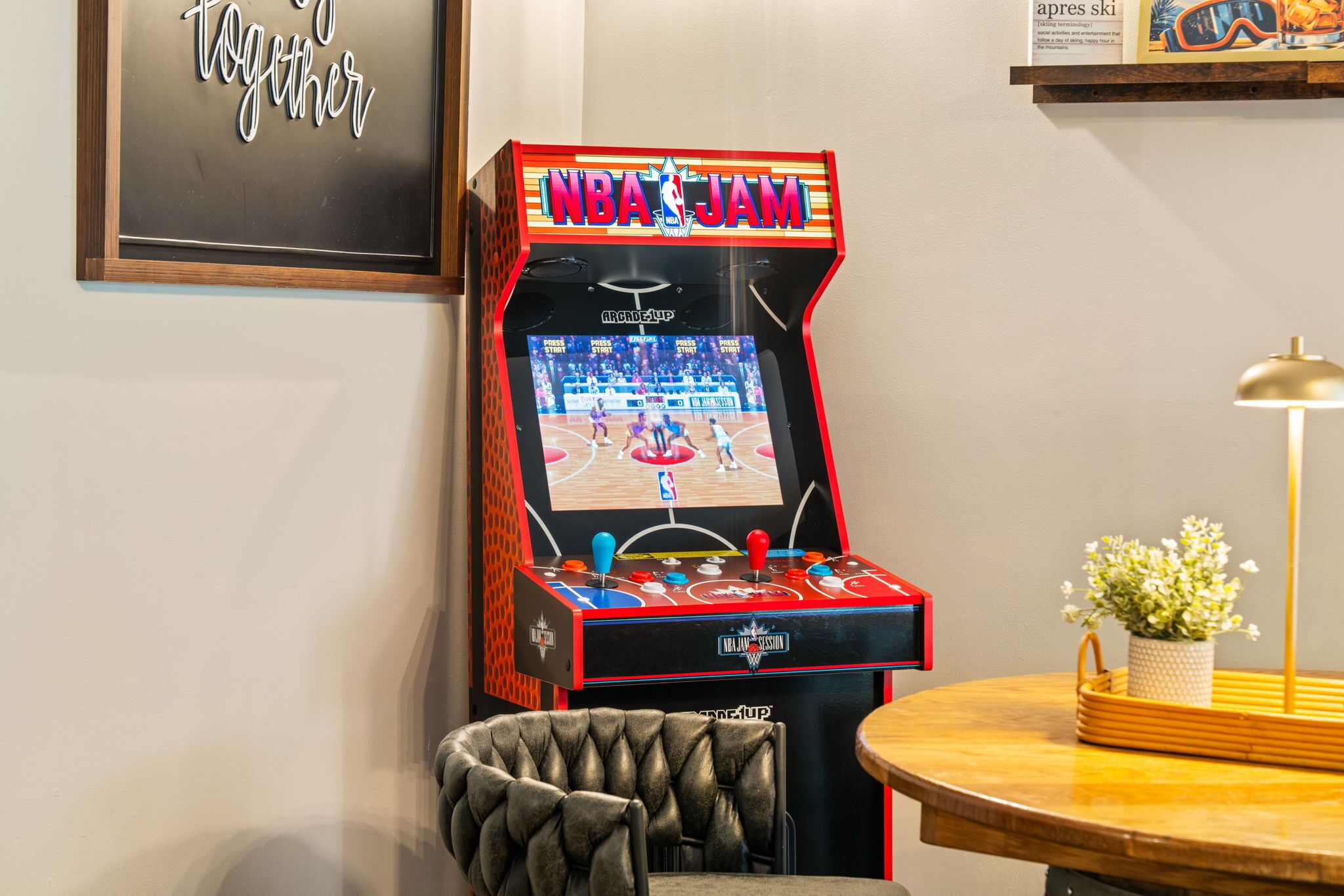 Une borne d’arcade avec un jeu de basket-ball, un fauteuil capitonné, une petite table avec une lampe et un panneau encadré accroché au mur.