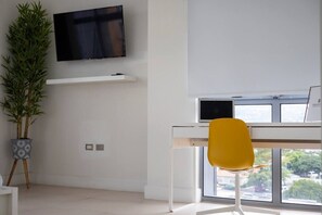 Ein modernes Zimmer mit Wand-Fernseher, Schreibtisch inklusive Computer, ein gelber Sessel und ein großes Fenster mit Blick auf Bäume.