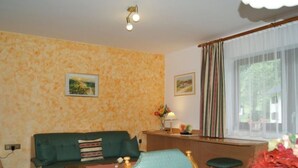 Ein Zimmer mit einem grünen Sofa, einem Holztisch, einem kleinen Hocker, einem Schreibtisch mit Lampe und einem Fenster mit Vorhängen.