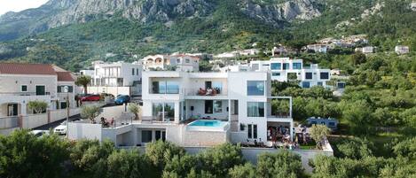 A modern multi-story building with a swimming pool, surrounded by lush greenery and mountains.