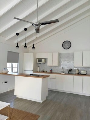 Une cuisine moderne avec des meubles blancs, un îlot central, un ventilateur de plafond, des luminaires suspendus, un four à micro-ondes et une horloge au mur.