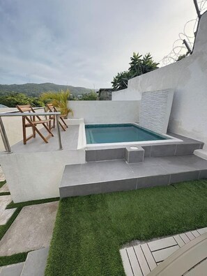 Une piscine avec un petit espace aménagé autour, entourée d’un mur blanc et offrant une vue sur des collines.