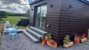 A modern, compact building with a dark exterior, large glass windows, and a small outdoor seating area with metal chairs and a table. - Hot Tub Getaway, Snowdon Views, Peaceful Stay (Llanfairpwllgwyngyll)