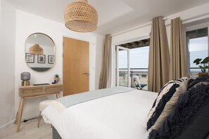 A bedroom with a bed, a bedside table, a mirror, and a view of the outside. - Trelantis, 5 Gwelva - Sea view, 250m to beach, Pets welcome (Newquay)