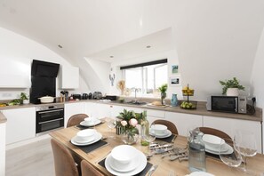 A modern kitchen with a dining area, equipped with a stove, oven, and microwave. - Trelantis, 5 Gwelva - Sea view, 250m to beach, Pets welcome (Newquay)