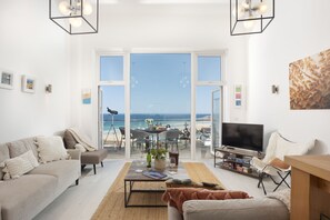A living room with a TV, sofa, and a balcony with a view of the sea. - Trelantis, 5 Gwelva - Sea view, 250m to beach, Pets welcome (Newquay)