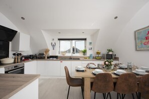 Eine moderne Küche mit weißen Schränken, ein Essbereich mit einem Holztisch und Stühlen sowie ein Ausblick ins Freie durch ein großes Fenster.