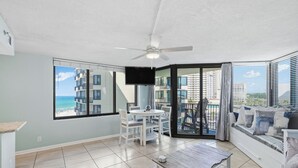 A balcony with a view of the ocean and buildings, a ceiling fan, a TV, and a dining area with chairs and a table. - Sunbird 612 East - Free Beach Chairs! (Panama City)