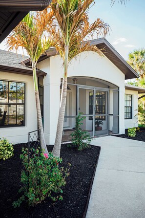 A modern house with a covered patio, a large window, and a well-maintained garden with a palm tree and shrubs. - The Gulf Access Oasis Heated Pool & SPA, Ramp/Dock (Port Charlotte)