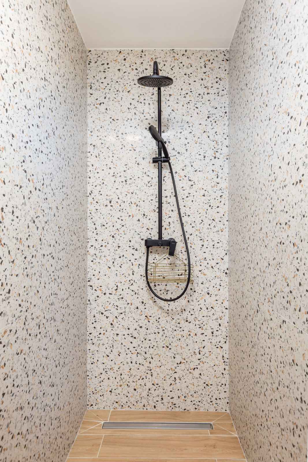 A shower with a handheld showerhead and a rain shower attachment, set against a speckled wall with a wooden floor.