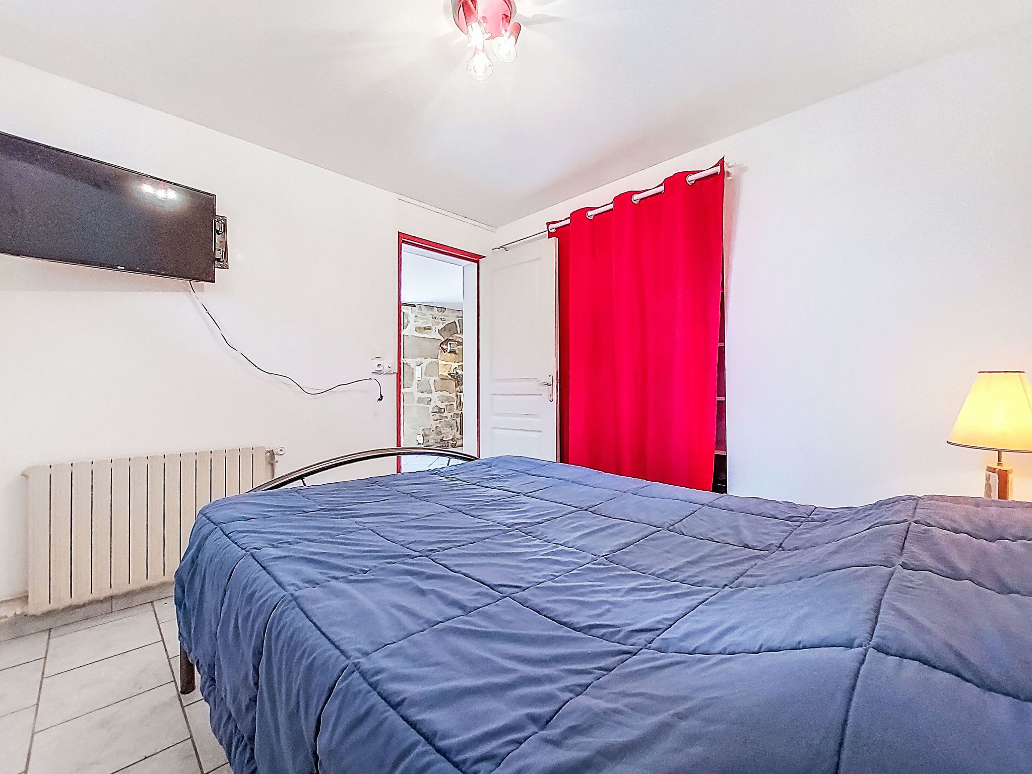 A bedroom with a bed, a TV mounted on the wall, a red curtain, and a lamp.