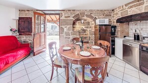 Une cuisine chaleureuse aux murs de pierre, une table à manger dressée pour quatre personnes, un canapé rouge et une vue dégagée sur l’extérieur grâce à une porte ouverte.