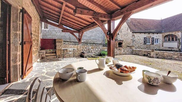 Un bâtiment en pierre avec un toit en bois, un mur en pierre et une table en pierre recouverte d’une nappe blanche, avec des bols et une assiette de nourriture.
