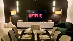 A dining area with a table set for six, a sofa, a TV displaying 'NETFLIX,' and a decorative wall.