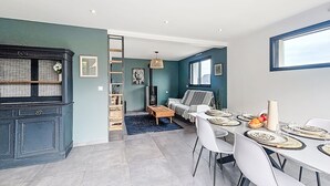 Et moderne køkken med indbygget komfur, et spiseområde med et langt bord og stole, og en opholdsstue med sofa og fjernsyn.