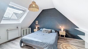 Et soveværelse med skråvæg, ovenlys, en seng med gråt sengetæppe, to sengeborde med lamper og en vævet måtte.