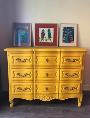 Un aparador amarillo con tiradores ornamentados y seis cajones, coronado por tres obras de arte enmarcadas.