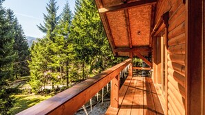 Um deck de madeira com guarda-corpo, um prédio de madeira com telhado inclinado e uma área verde ao fundo.